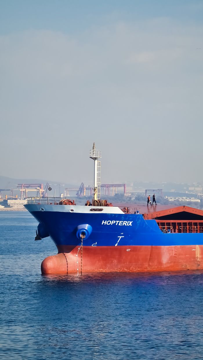 gallery-4 Close-up of a blue cargo ship on calm waters with an industrial port in the background.