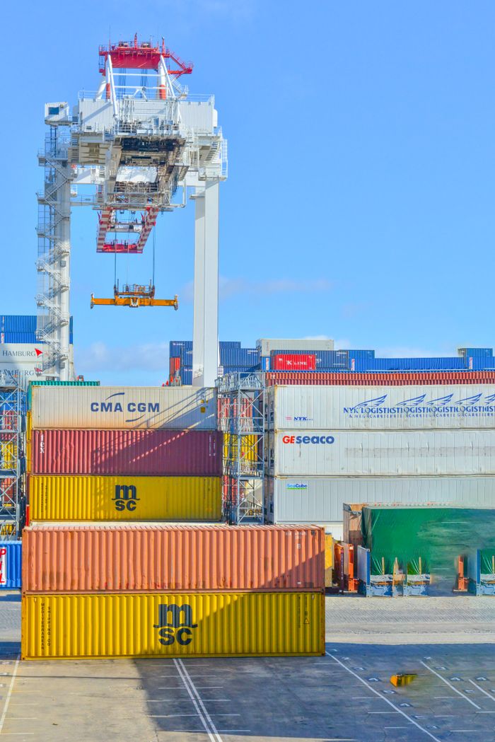 our-services-1 Stacked cargo containers in a sunny port industrial setting with crane.