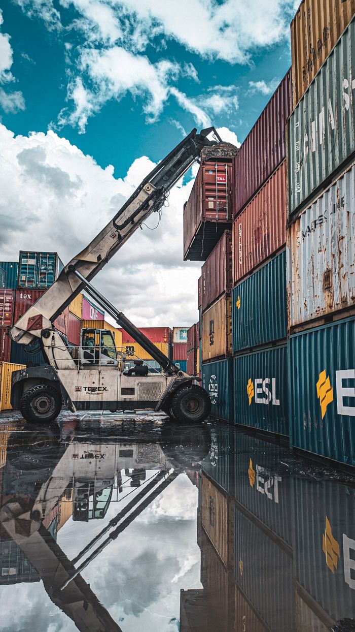 gallery-1 A crane loader stacking colorful shipping containers at a port with reflection in water.